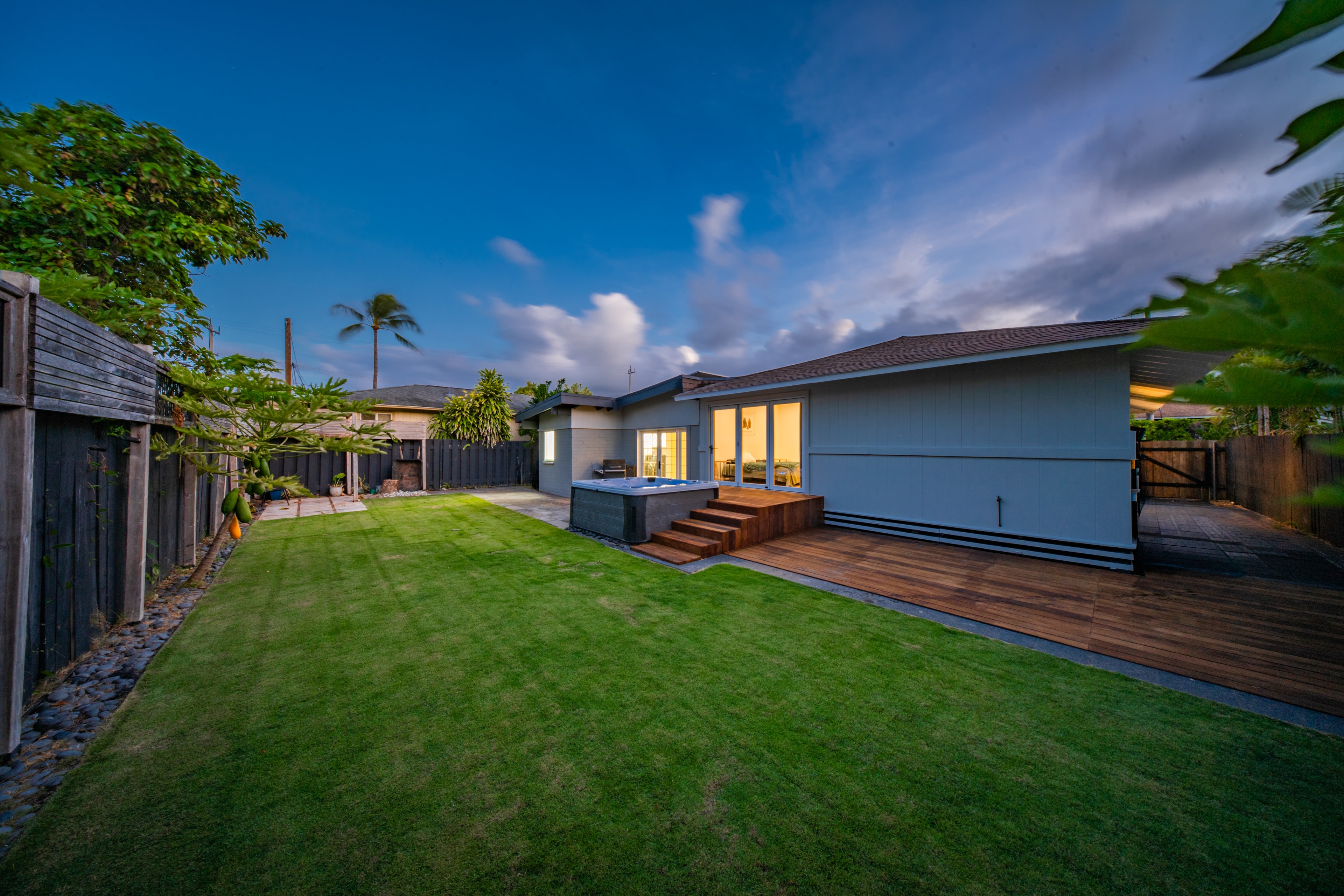 Backyard at dusk — wide lawn, wood deck, hot tub, and palm trees against a deep blue sky