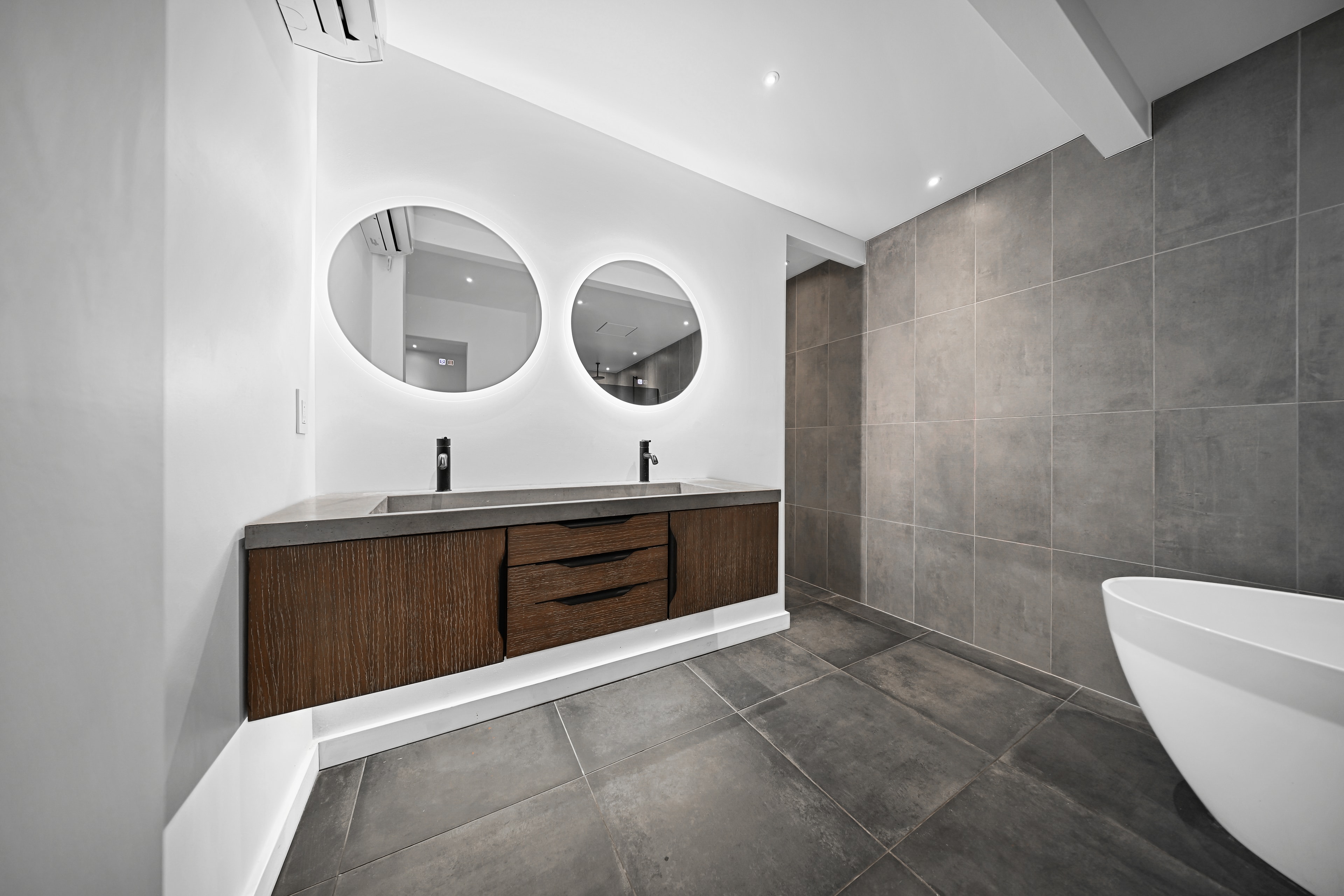 Primary bath wide view: dual backlit round mirrors, floating wood vanity, concrete sinks, large-format tile, and freestanding tub