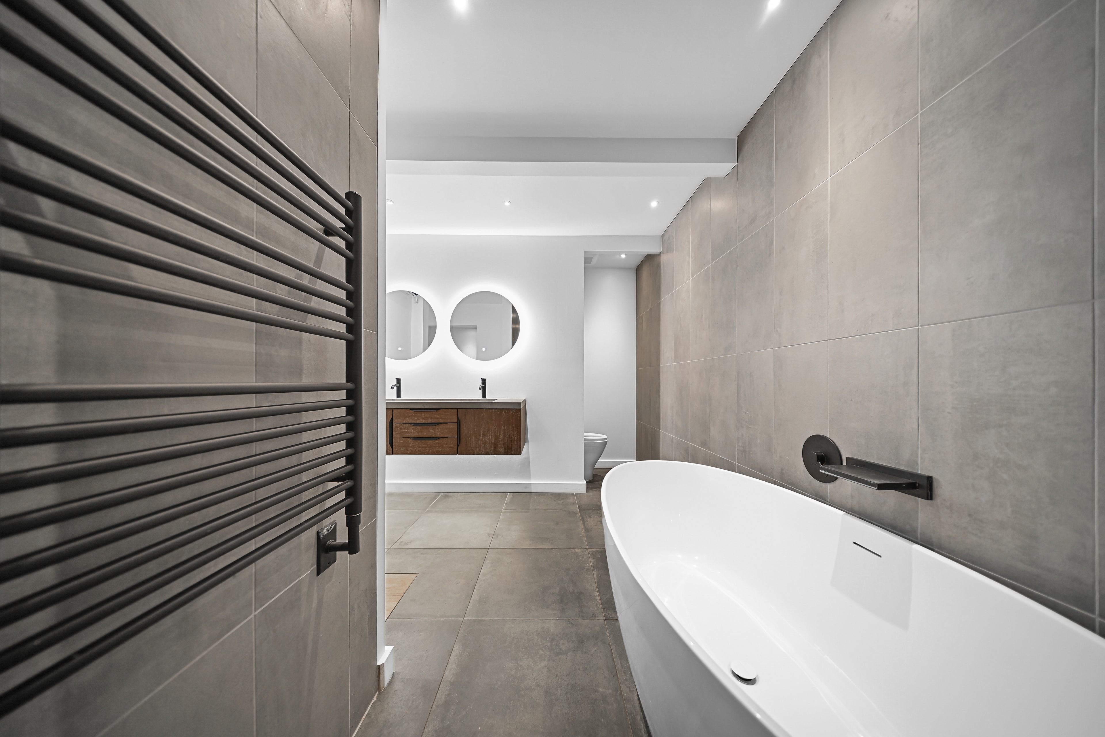 Primary bath with matte black heated towel rack, freestanding soaking tub, dual backlit mirrors, and large-format concrete tile