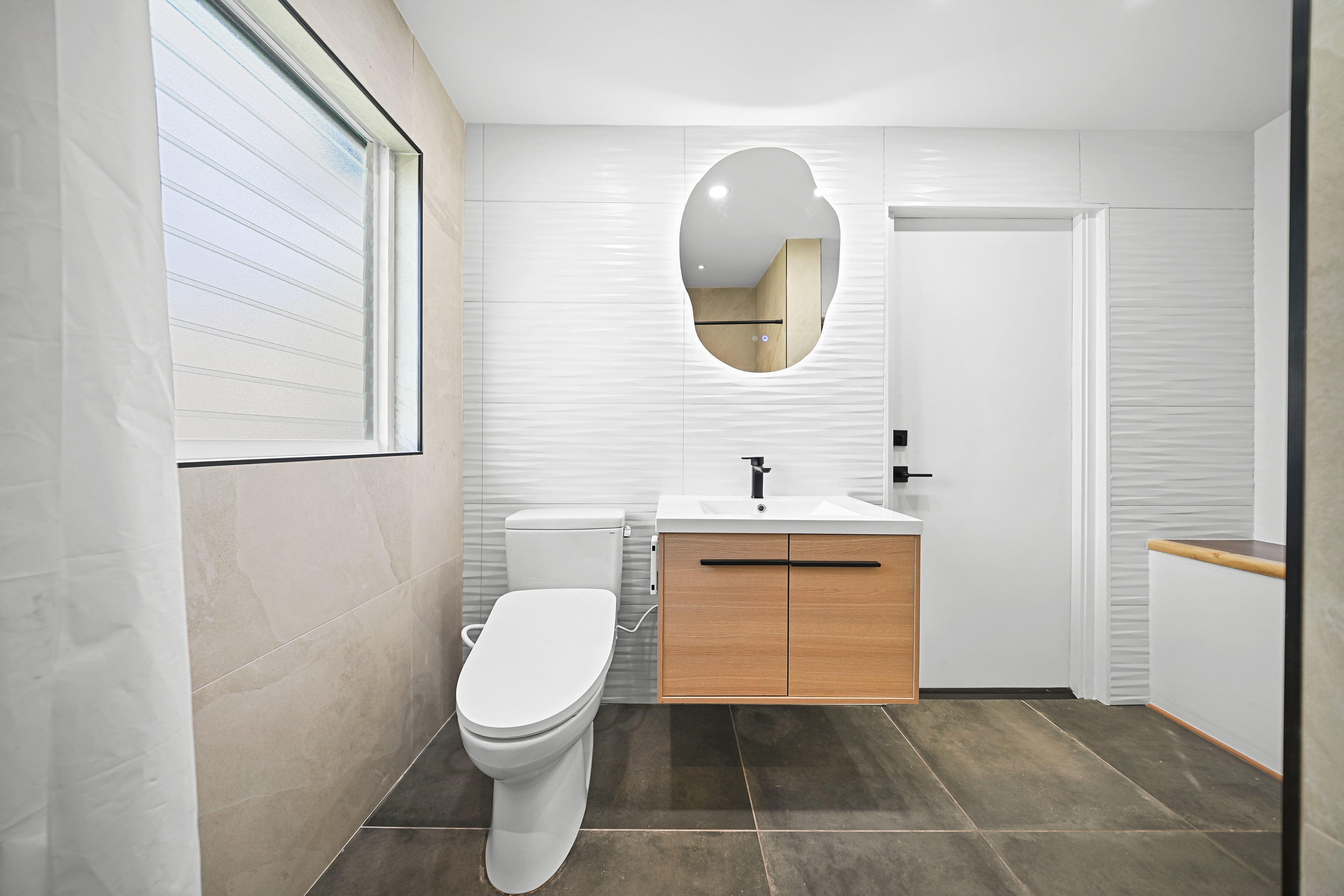 Bathroom with floating wood vanity, sculptural backlit mirror, wave-tile accent wall, and dark tile floor