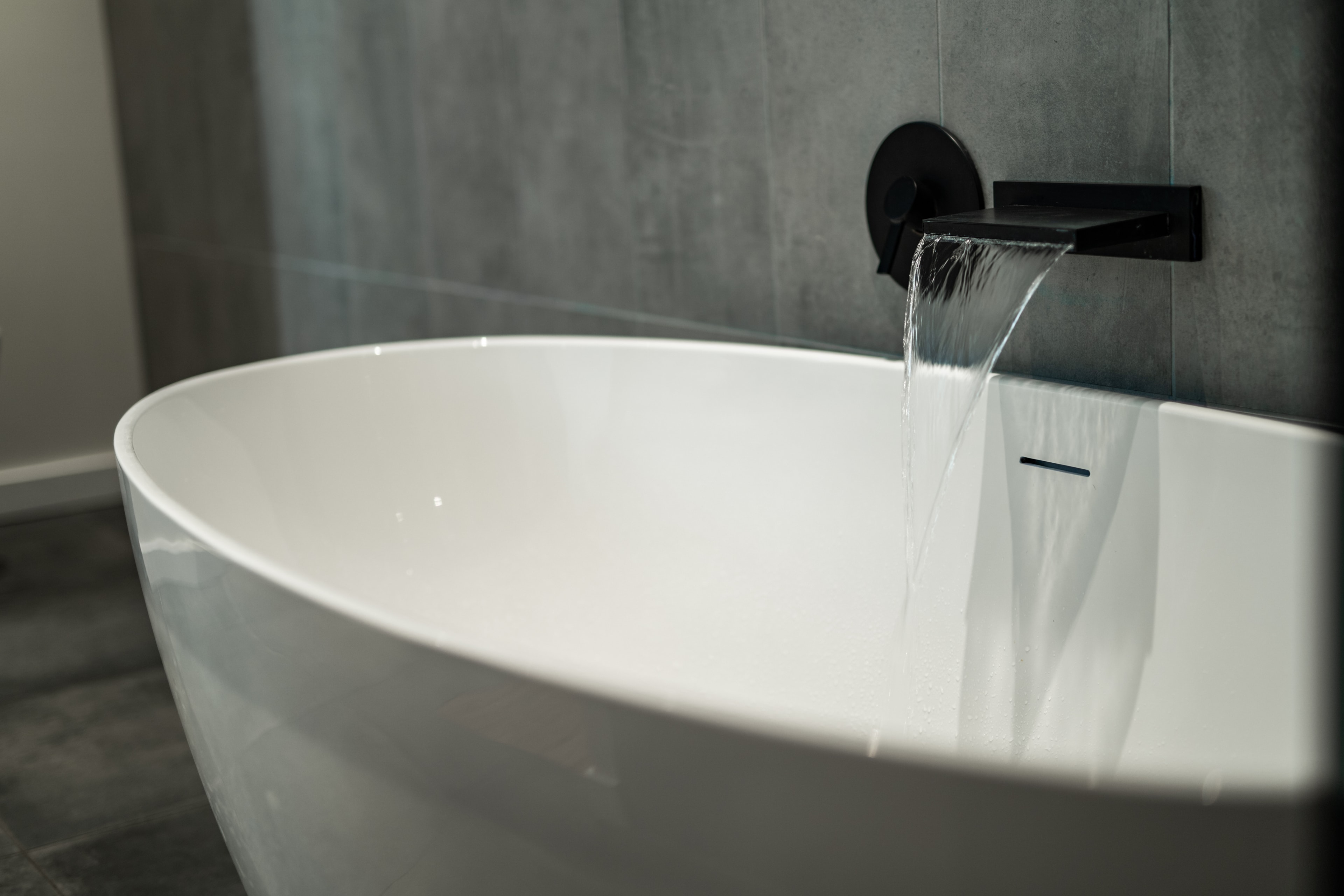 Freestanding soaking tub with wall-mounted waterfall faucet and dark tile surround