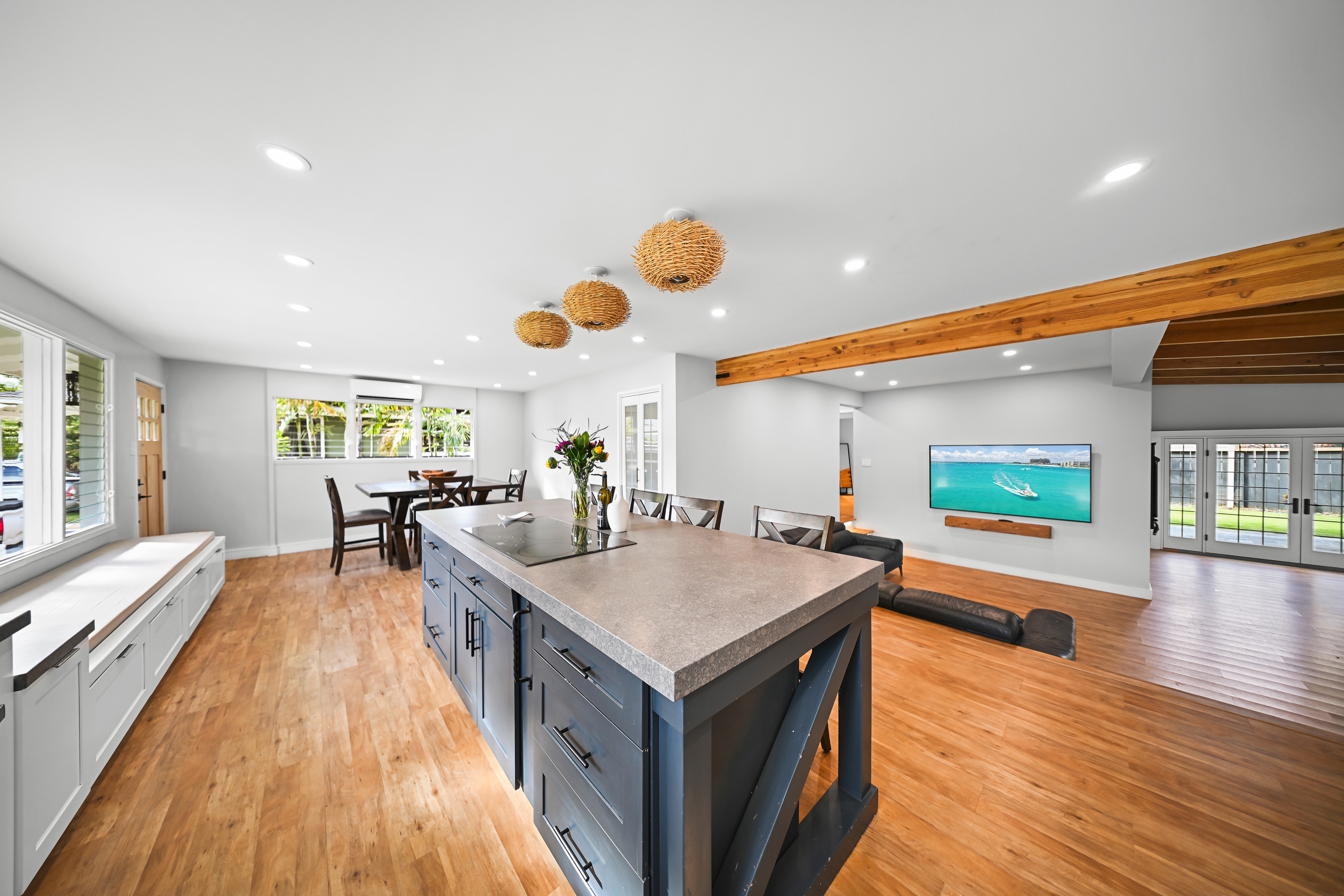 Great room wide shot: dark island, woven pendants, exposed beam, large TV wall, and French doors to backyard