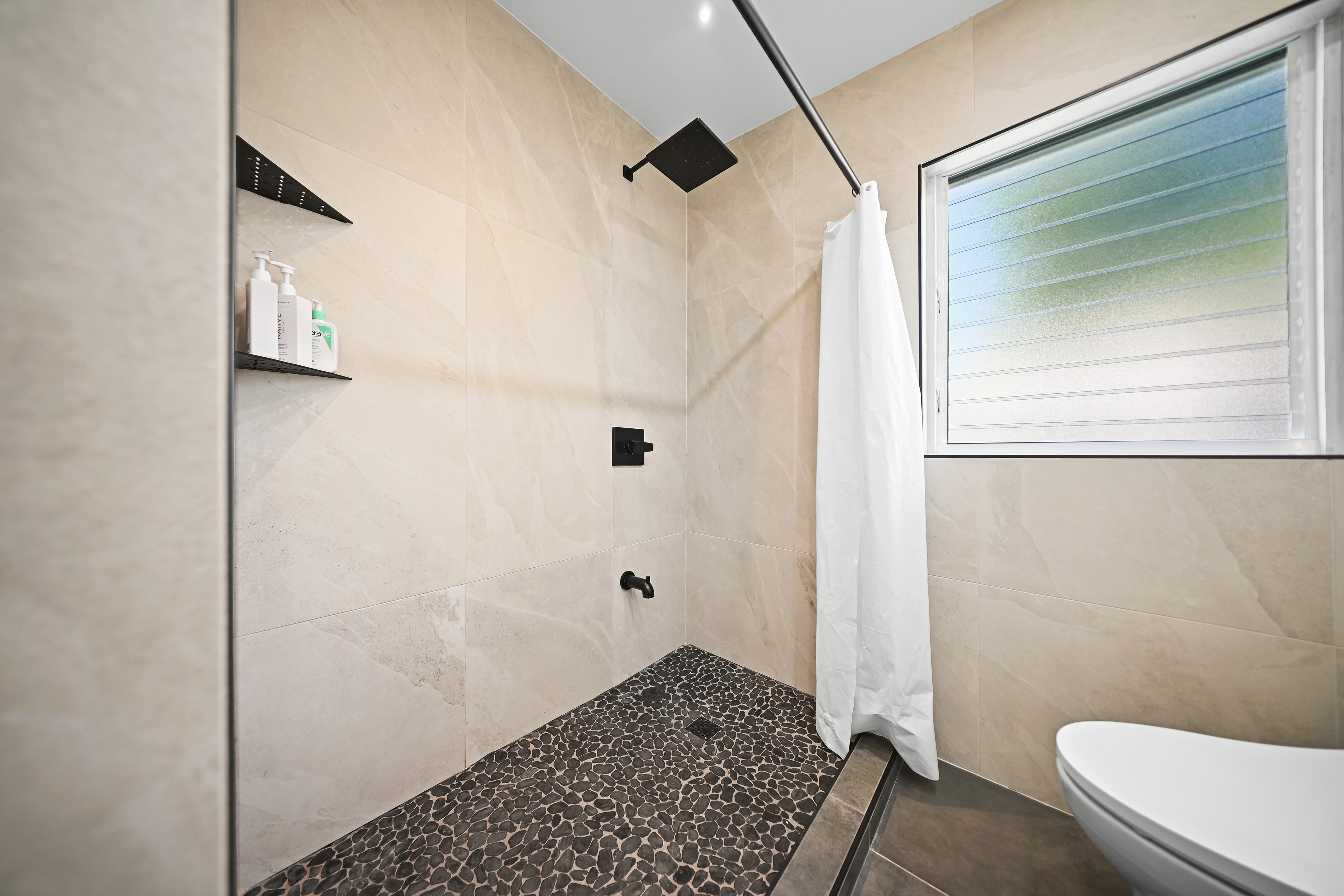 Walk-in shower with large beige tile, pebble stone floor, matte black rain head, and louvered window